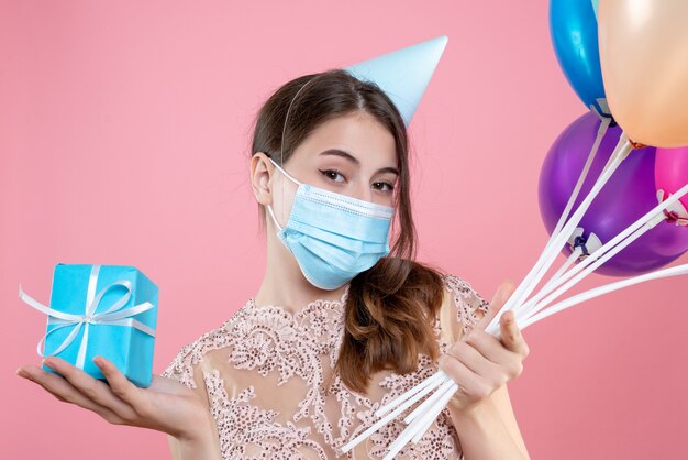 Closeup front view cute party girl with crown and mask holding balloons and gift