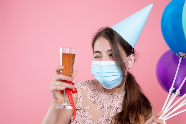 Closeup front view cute party girl with crown and mask holding balloons toasting