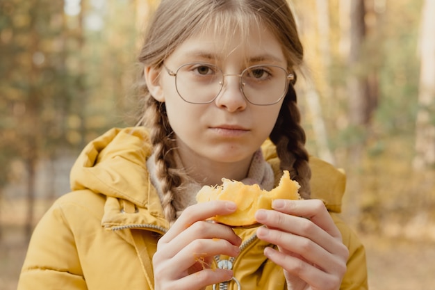 안경과 노란색 재킷을 입은 근접 촬영 소녀는 호박을 먹습니다. 야외 피크닉. 할로윈을 준비하는 작은 도우미 프리미엄 사진
