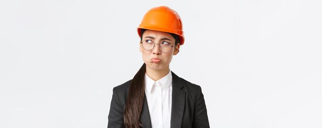 Closeup of gloomy upset asian female engineer in safety helmet and business suit complaining looking