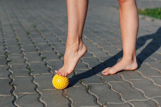 Closeup of graceful bare feet of young woman standing on tiptoes on spiked massage ball on paving sl...