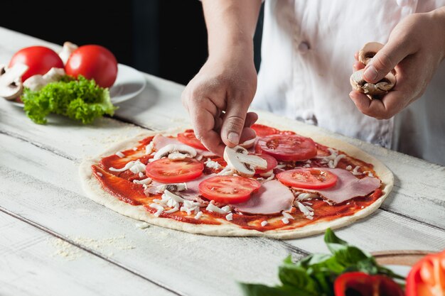 Крупным планом рука шеф-повара пекаря в белой форме, делая пиццу на кухне