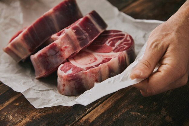 Closeup hand holds white craft paper with three raw great leg steaks with bone on it, everything on old wooden brushed table