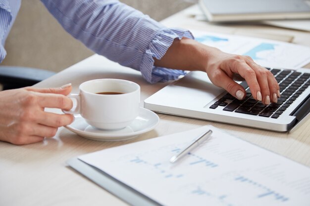 Closeup of hands of businesswoman keyboarding on laptop and holding a cup of tea