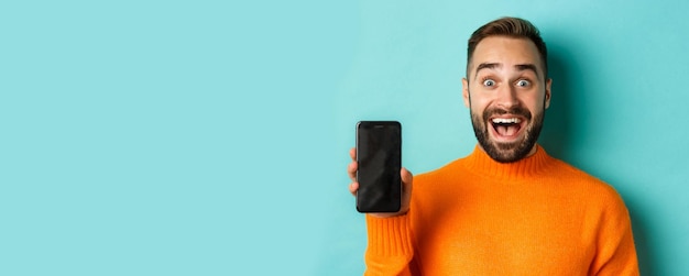 Крупный план красивого бородатого парня в оранжевом свитере, показывающего экран смартфона и улыбающегося, показывающего выпускной вечер