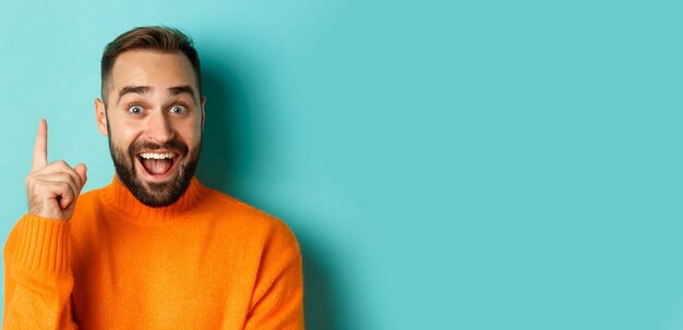 Closeup of handsome man having an idea raising finger up and smiling excited standing in orange swea