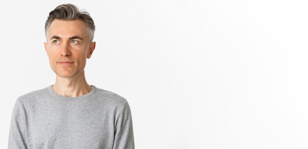 흰색 위에 서있는 왼쪽 위 모서리를 바라보는 회색 스웨터를 입은 잘생긴 중년 남자의 근접 촬영