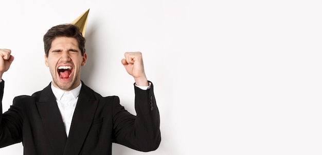 Free photo closeup of happy goodlooking man wearing party hat and suit raising hands up and rejoicing celebrati
