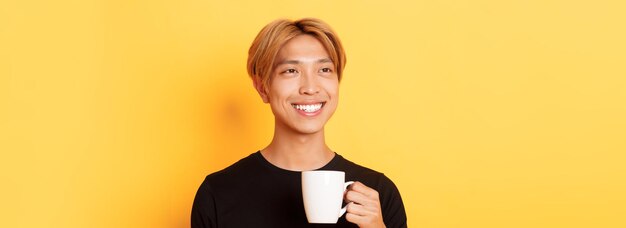 Closeup of happy handsome young asian guy with blond hair looking dreamy and smiling while drinking