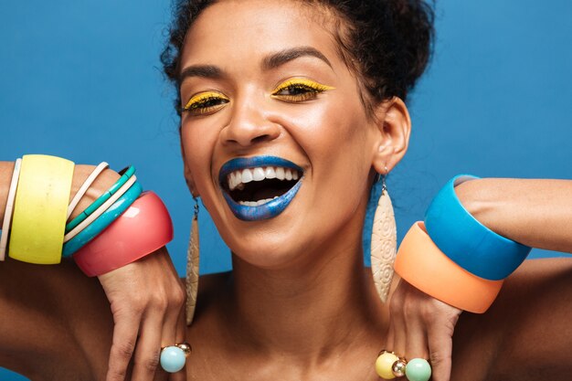 Closeup happy mulatto woman with colorful makeup laughing and showing beautiful adornment on her arms, isolated over blue