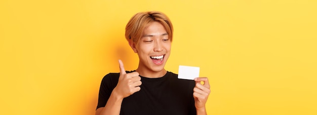 Primo piano del ragazzo bello asiatico felice e soddisfatto che mostra la carta di credito e thumbsup nello smili di approvazione