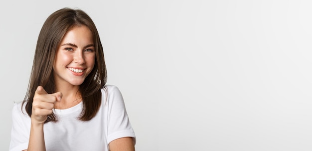 Closeup of happy smiling brunette girl pointing at camera standing white background