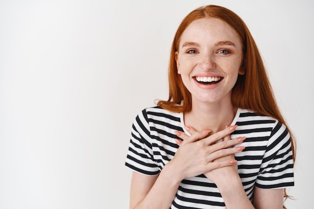 Foto gratuita primo piano di felice giovane donna con lentiggini e capelli rossi che si tengono per mano sul petto e sorridente ringraziandoti in piedi sinceramente contro uno sfondo bianco