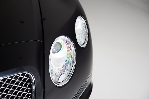 Closeup of the headlight of a black luxury car under the lights against a grey background