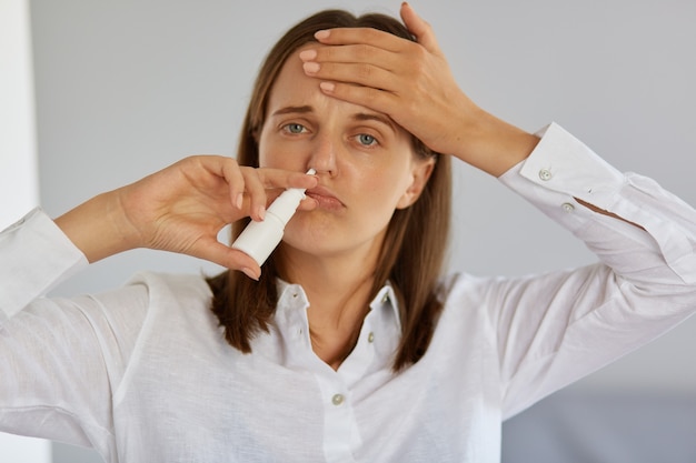 Closeup of ill young adult woman using nasal spray, suffering from runny nose and terrible headache, looking at camera, touching her forehead, having flu and high temperature.