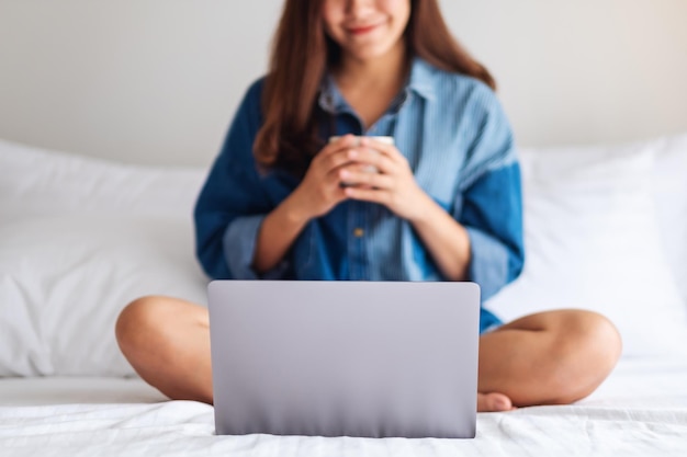 집에서 침대에 앉아서 커피를 마시는 동안 화상 통화를 위해 노트북 컴퓨터를 사용하는 아름다운 아시아 여성의 클로즈업 이미지