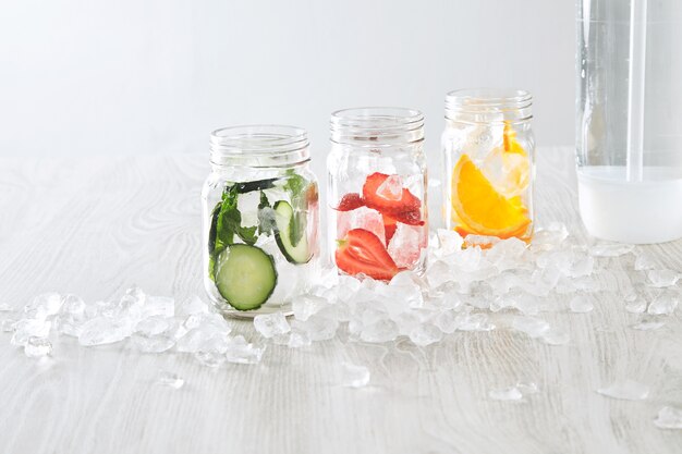 Closeup jars with ice and various fillings orange,strawberry,cucumber and mint prepared to make fresh homemade lemonade with sparkling water