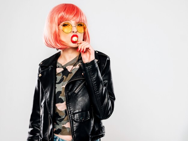 Closeup lips of young beautiful hipster bad girl in trendy black leather jacket and earring in her nose.Sexy carefree smiling woman posing in studio in pink wig.Positive model licking round candy