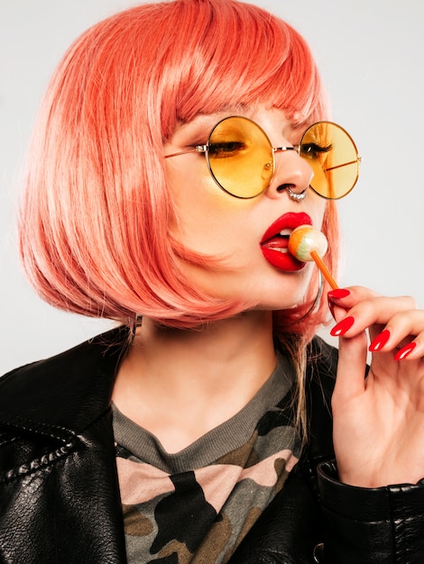 Closeup lips of young beautiful hipster bad girl in trendy black leather jacket and earring in her nose.Sexy carefree smiling woman posing in studio in pink wig.Positive model licking round candy