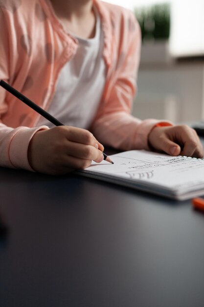 Closeup of little schoolchild writing mathematics homework on notebook during classroom lesson sitting at desk table in living room. Concept of home schooling, distance learning, online education