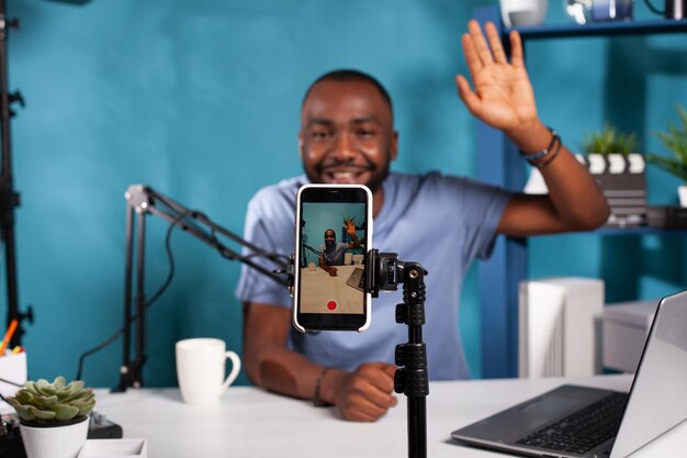 Closeup of live vlog setup with smarthone on stand filming content creator waving hand to followers in online morning show. Selective focus on vlogger saying hello at audience sitting at desk.