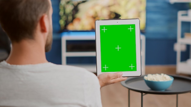 Closeup of man holding vertical digital tablet with green screen in online conference or group video call in home living room. Person using touchscreen device with chroma key watching webinar.