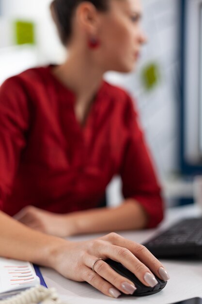 Крупный план пальцев менеджера с кольцом, щелкающим за столом в стартовом офисе. Сотрудник прокручивает содержимое на настольном компьютере. Рука предпринимателя в красной рубашке с помощью беспроводной компьютерной мыши.