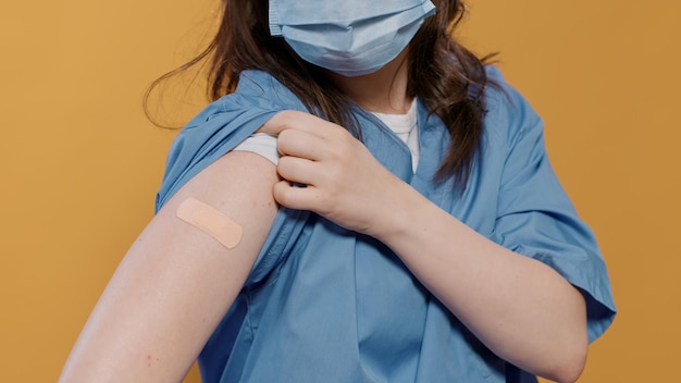 Closeup of medical doctor wearing surgical mask lifting sleeve and showing band aid after covid or flu vaccine in studio. Detail view of medic in uniform revealing bandage covering immunization spot.