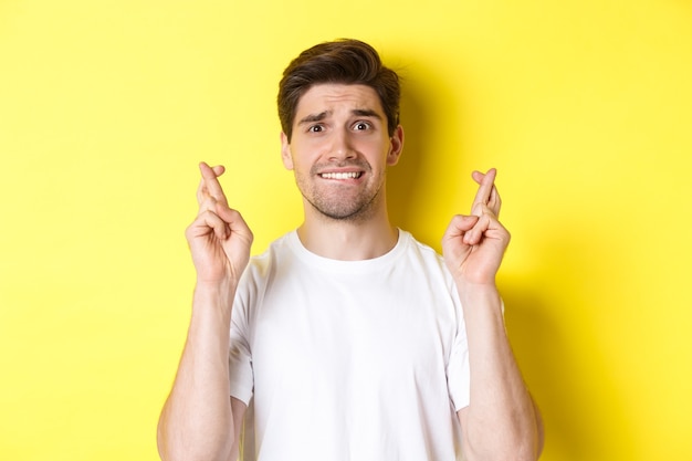 Closeup of nervous man making wish holding fingers crossed and biting lip worried standing over yell...