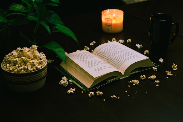 Closeup of an open book and a bowl of popcorn on the table with a lit candle and a cup of coffee