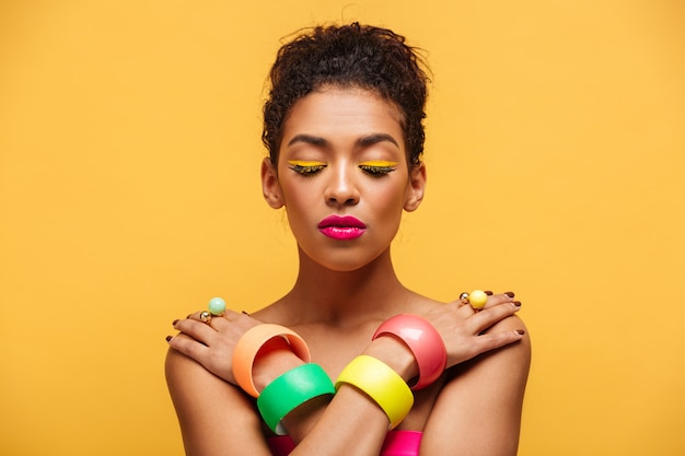 Closeup peaceful mulatto woman with closed eyes and pink lipstick posing on camera with crossed hands on shoulders, over yellow wall