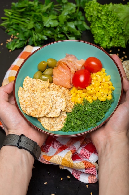 Closeup of a person holding a bowl of salad with salmon, crackers and vegetables under the lights