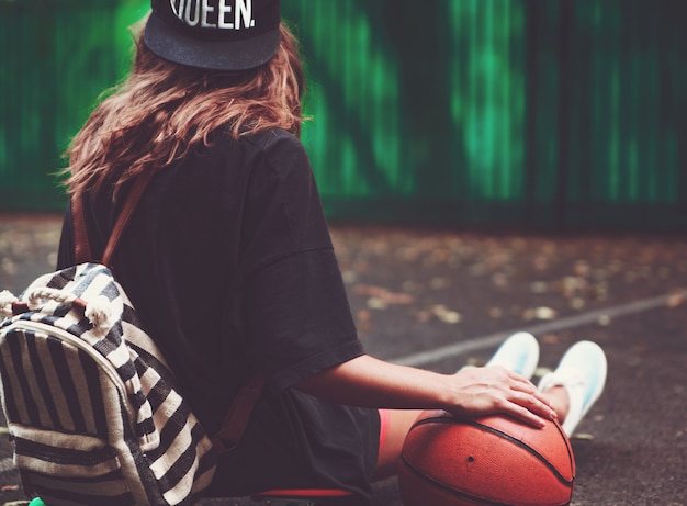 Closeup photo basketball ball with girl sitting on plastic orange penny shortboard on asphalt