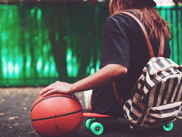 Closeup photo basketball ball with girl sitting on plastic orange penny shortboard on asphalt