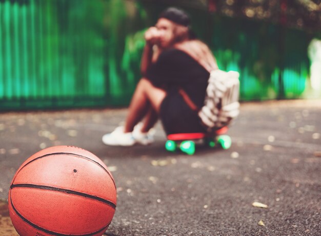 Closeup photo basketball ball with girl sitting on plastic orange penny shortboard on asphalt