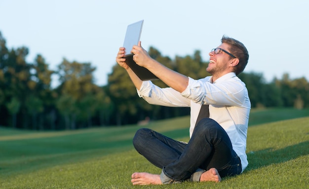 休憩中に自分撮りをしているフリーランサーのクローズアップ写真緑の芝生に座って写真を友達に送っている眼鏡をかけた男
