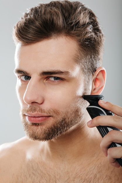 Closeup picture of good-looking adult guy grooming his face with shaving his cheek using electric shaver over grey wall