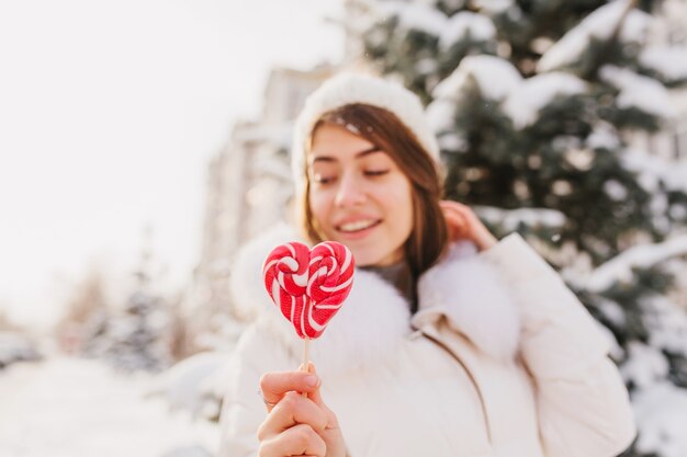 晴れた朝に雪でいっぱいの路上でゾッと手冬女性のクローズアップピンクハートロリポップ。白いニット帽、楽しんで。おいしい、甘い生活、冬休み。