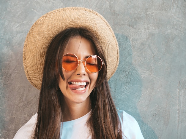 Portraif del primo piano di giovane bello sguardo sorridente della donna. ragazza alla moda in abiti casual e cappello estivo. femmina positiva che fa fronte divertente e che mostra lingua