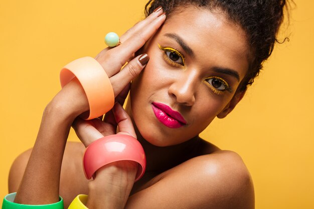 Closeup portrait of adorable mulatto woman with yellow eyelids and pink lips looking on camera holding hands at face, isolated over wall