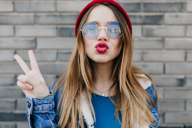 Closeup portrait of amazing woman with kissing face expression standing in front of brick wall Outdoor photo of cheerful young lady in glasses and denim jacket posing on the street