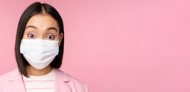Крупным планом портрет азиатской бизнес-леди в медицинской маске, выглядящей удивленной, стоящей в костюме на розовом фоне