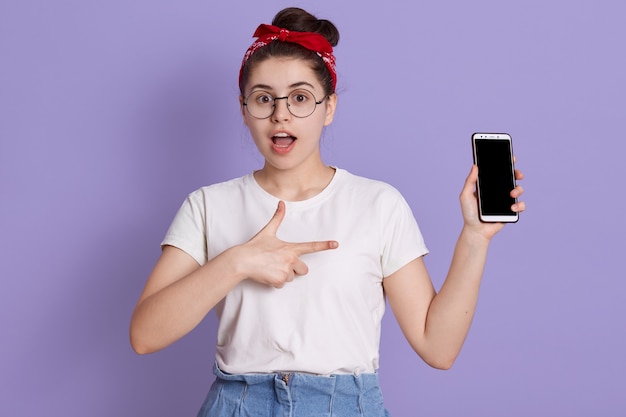 Ritratto del primo piano della donna stupita che posa con la bocca aperta e che tiene il telefono astuto moderno con lo schermo in bianco