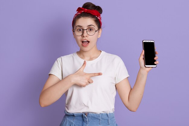 驚いた女性が開いた口でポーズと空白の画面を持つ現代のスマートフォンを押しのポートレート、クローズアップ