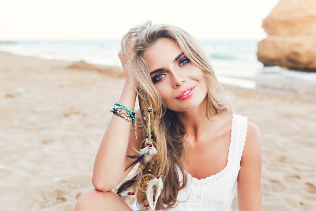 Портрет крупного плана привлекательной белокурой девушки с длинными волосами и голубыми глазами, сидя на пляже. Она смотрит в камеру.
