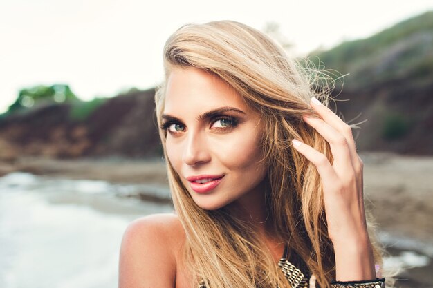 Closeup portrait of attractive blonde girl with long hair posing on rocky beach. She is touching hair and looking to the camera.