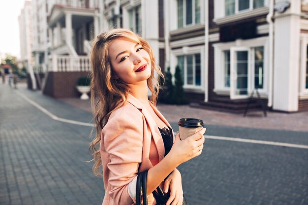 Closeup portrait  attractive model with vinous lips walking with coffee in coral jacket on street.