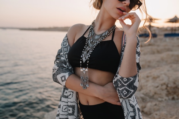 Ritratto del primo piano della ragazza attraente che toglie gli occhiali da sole alla moda, sul tramonto, sulla spiaggia con sguardo sensuale. indossa top nero alla moda, collana, cardigan con ornamenti. resort, vacanza, vacanza.