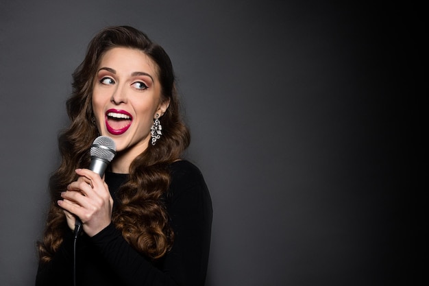 Closeup portrait of beautiful lady with red lips singing in studio. professional woman in black dress looking away while posing over black background.