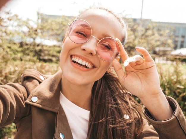 Closeup portrait of beautiful smiling brunette girl in summer hipster jacket  Model taking selfie on smartphone Woman making photos in warm sunny day in the street in sunglasses